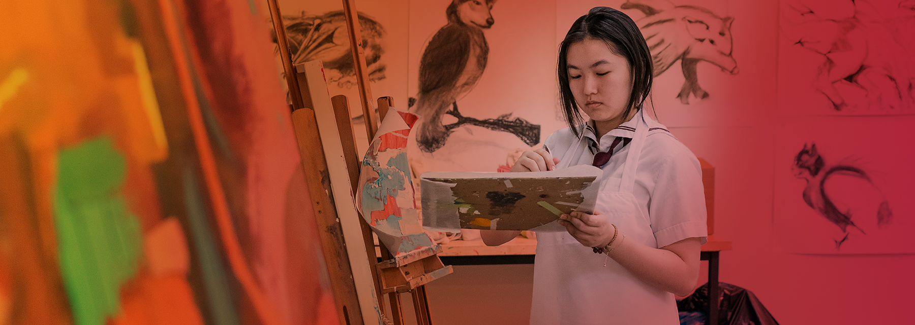 A senior school student wearing an art smock and holding a painting palette stands in front of a large colourful canvas.